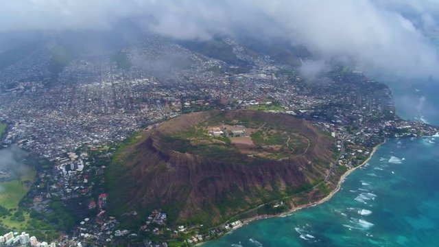 High altitude aerial shot of Diamond Head crater and beach, moving offshore through white clouds. Waves breaking on the reef and sandy beach far below. 