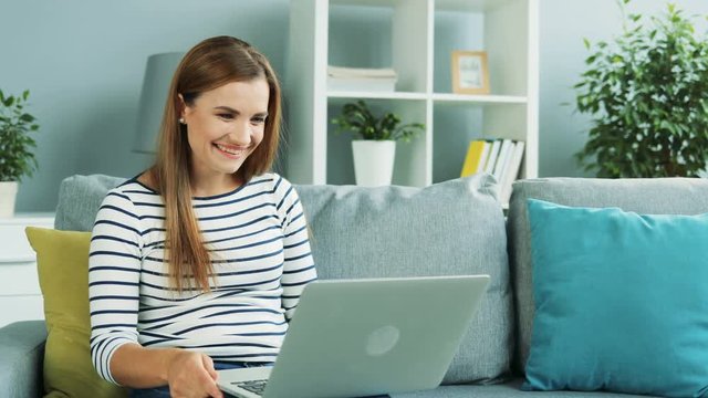 Attractive woman sitting on the couch with pillows and having video chat on the laptop computer in the cozy room. Inside