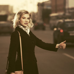 Fototapeta premium Fashion blond woman in black coat hailing a taxi cab walking in city street