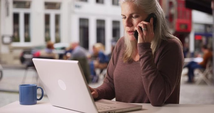 Busy Older White Woman Works From Laptop And Makes Phone Call At Cafe