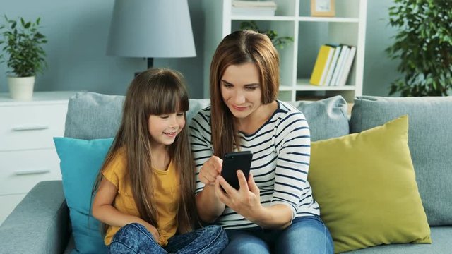 Cute little girl sitting with her young mother on th couch in the living room and scrolling photos on the black smartphone. Inside