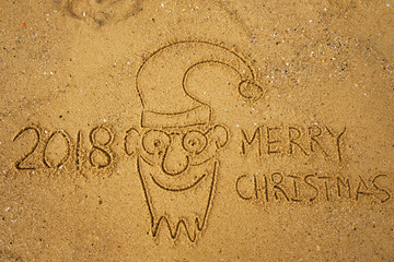 Handwriting words MERRY CHRISTMAS 2018 on sand of beach and funny face of santa claus. Concept of travel in winter