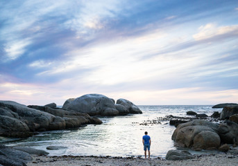 A man standing on the beach at the edge of the ocean watching the sunset through the clouds in Cape Town in summer