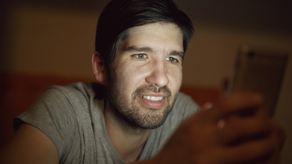 Closeup of Young smiling man using smartphone for surfing social media lying in bed at home at night