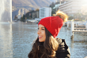 Happy winter woman with red wool hat and pompon with the city lake on the background - obrazy, fototapety, plakaty