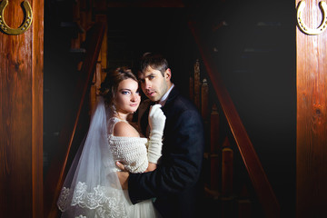 The groom in a black coat and the bride in a white wedding dress hugging in the background is a wooden ladder in a dark room