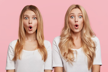 Two surprised blonde sisters stand next to each other, keep mouth opened and stare at camera,...