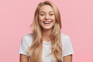 Portrait of young blonde positive female with cheerful expression, dressed in casual white t shirt, rejoices to recieve good job offer, isolated over pink background. Beautiful woman indoor. - obrazy, fototapety, plakaty