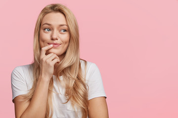 Pretty lovely blonde female with pure healthy skin dressed casually looks with dreamy expression aside, remembers pleasant moments, isolated over pink background, copy space for your advertisment