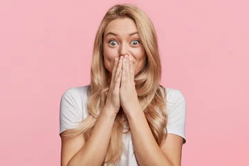 Deurstickers Beautiful excited blonde female covers eyes with happiness, doesn`t expect to recieve pleasant surprise from close friend, being thankful and pleasantly shocked, isolated over pink background © Wayhome Studio