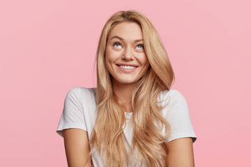 Cheerful blonde female looks upwards, has shining smile, thoughtful expression, dressed in casual...