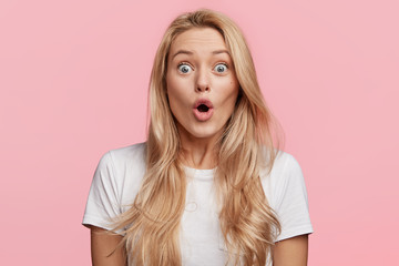 Pretty blonde woman stares at camera, keeps mouth widely opened, dressed in casual t shirt, expresses great surprisment, feels shocked about sales in web sites, wines in online contest, wonders news