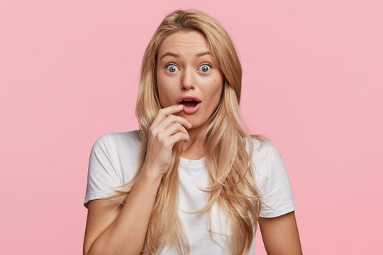 Surprised positive female with blue eyes popped out, opens mouth widely in surprisment as hears news from interlocutor, straes at camera, expresses shock and disbelief, isolated on pink wall