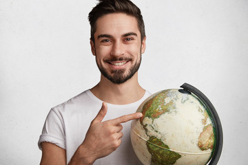 Handsome cheerful young active bearded traveller holds globe, chooses destination where to go,...