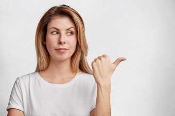 Young pretty housewife dressed in casual white t shhirt, spends time at home, indicates with thumb...