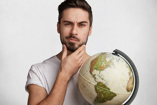 Portrait of thoughtful bearded man tourist with globe chooses country to travel, has serious and pensive look, isolated over white concrete background. Clever intelligent male teacher of geography