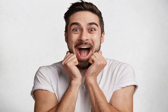 Excited joyful bearded male looks with amazement at camera, being overjoyed as sees unexpected surprise, isolated over white concrete background. People, facial expressions and happiness concept