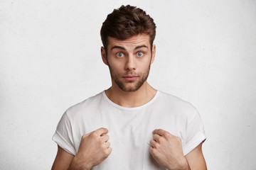 Portrait of puzzled young man with blue eyes and stubble wears casual t shirt, points at himself looks in bewilderment as doesn`t understand something, isolated over white concrete background