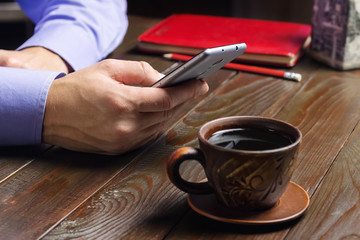 Fototapeta na wymiar Home office workplace, male hands hold a smartphone, cup of black coffee, notepad on dark wood working table
