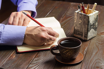 Male hand with red pencil writes in notepad, black coffee in brown cup on dark wood working table, working or study concept