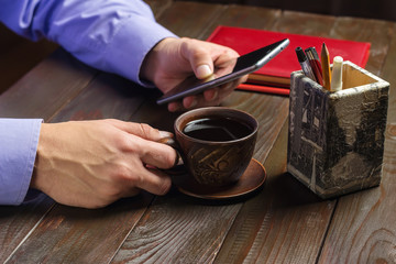 Fototapeta na wymiar Home office workplace, male hands hold a cup of black coffee and smartphone, notepad on dark wood working table, a coffee break time