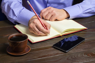 Man in blue shirt with red pencil in hand writes in notepad, cup of black coffee and smartphone on dark wood working table, working or study concept