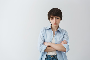 Portrait of good-looking masculine girl with short dark hair and brown big eyes, crossing hands, having aggressive and confident look.