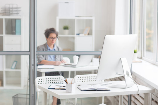 Computer monitor, open notebook, picture frame, document and pencil-box on desk by transparent wall and manager working behind it