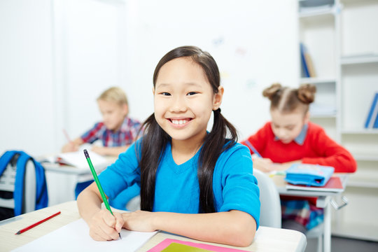 Happy Asian schoolgirl making notes or sketching at lesson and looking at camera