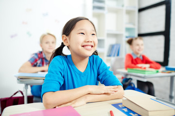 Cute smiling schoolgirl listening to teacher while sitting by desk at lesson