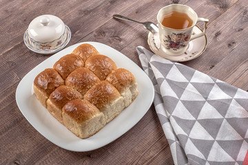 White bread in glass plate, spoon, napkin, butter dish and cup of tea on wooden table