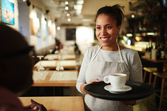 Young hospitable waitress bringing cup of fresh coffee on tray for her client