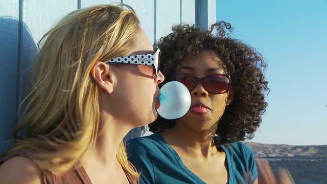 Two women friends in sunglasses blowing bubblegum at the beach