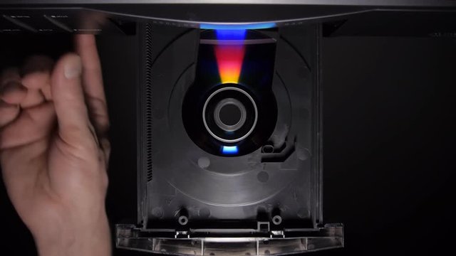 The hand puts the compact disk in the DVD player and after play get it back on black background. Bottom view