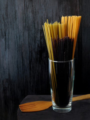 Different sorts of spaghetti in a glass decorated with basil leaves on a dark wooden background