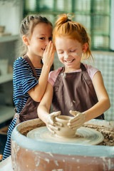 pottery workshop - two girls sculpts from clay. one child whispers from her friend
