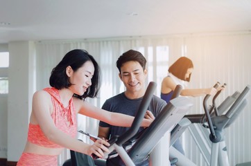 asian young woman slim body exercising on bicycle machine with young man personal trainer in fitness gym, bodybuilder, healthy lifestyle, fitness, workout and sport training concept, soft focus