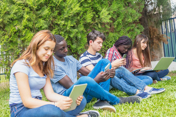 Teenagers of different races using tablets, smartphones and laptops in a city park