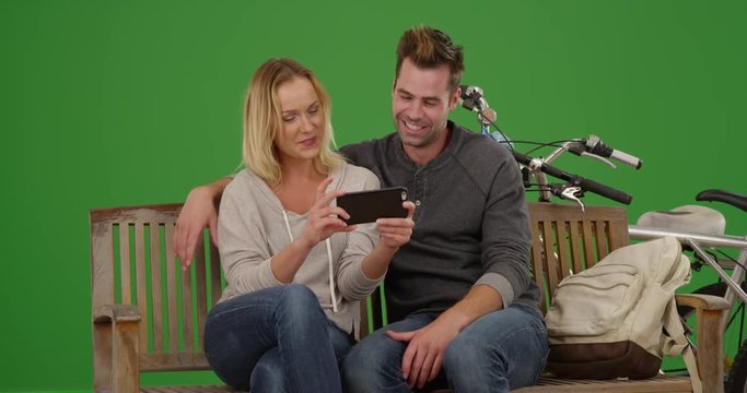 Millennial couple watching videos on smartphone together on a bench on green screen. On green screen to be keyed or composited. 