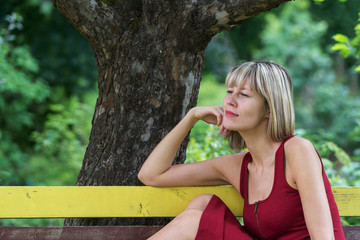 Naklejka premium Young blond woman in a red dress leaning sits on a wooden bench. .Businesswoman walking along the street and resting in the green city park on the beautiful sunny summer day.
