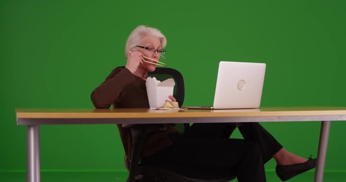 Mature white woman eating Chinese food while watching something on her laptop on greenscreen. Older woman using her laptop while eating on green screen to be keyed or composited. 
