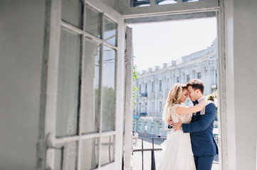 The bride and groom stand on the balcony and kissing. Enamored newlyweds embrace. Studio in white interior.