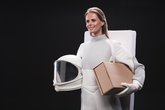 Ready for change. Pleasant optimistic spacewoman wearing protective suit is holding cardboard box and helmet. She is going to spaceship while is looking aside with smile. Isolated with copy space