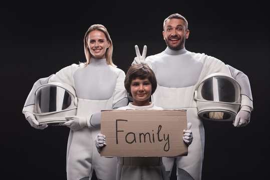 Family concept. Portrait of happy couple of cosmonauts are standing together with their son. Child is holding cardboard sign family while looking at camera with joy. Isolated background