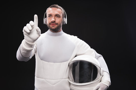 Choose best song. Portrait of pleasant bearded astronaut in headphones is standing in front of transparent screen and pressing on virtual button with concentration. He is holding helmet. Isolated