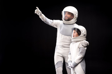 Look at this. Positive cosmonaut father is hugging his son while pointing on something while boy is feeling wonder. They are standing together wearing protective costume. Isolated with copy space