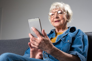 Portrait of old smiling lady looking at screen of mobile phone while sitting on couch