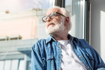 Morning relax. Calm pensioner looking outside while resting near window at living room. Copy space in left side