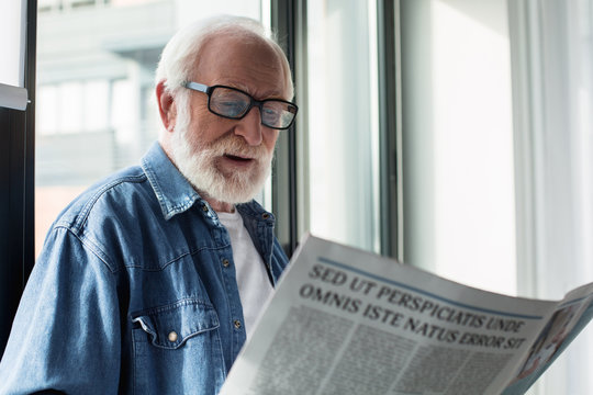 Portrait of old bearded pensioner enjoying newspaper at home. He is cheerful and interested