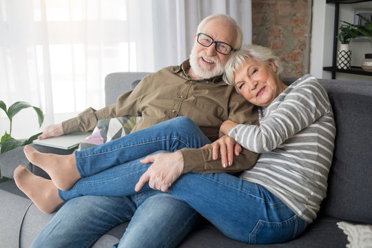 Happy rest. Portrait of delightful senior husband and wife relaxing on couch at home. Woman is tenderly cuddling man, while smiling male tightly hugging her legs